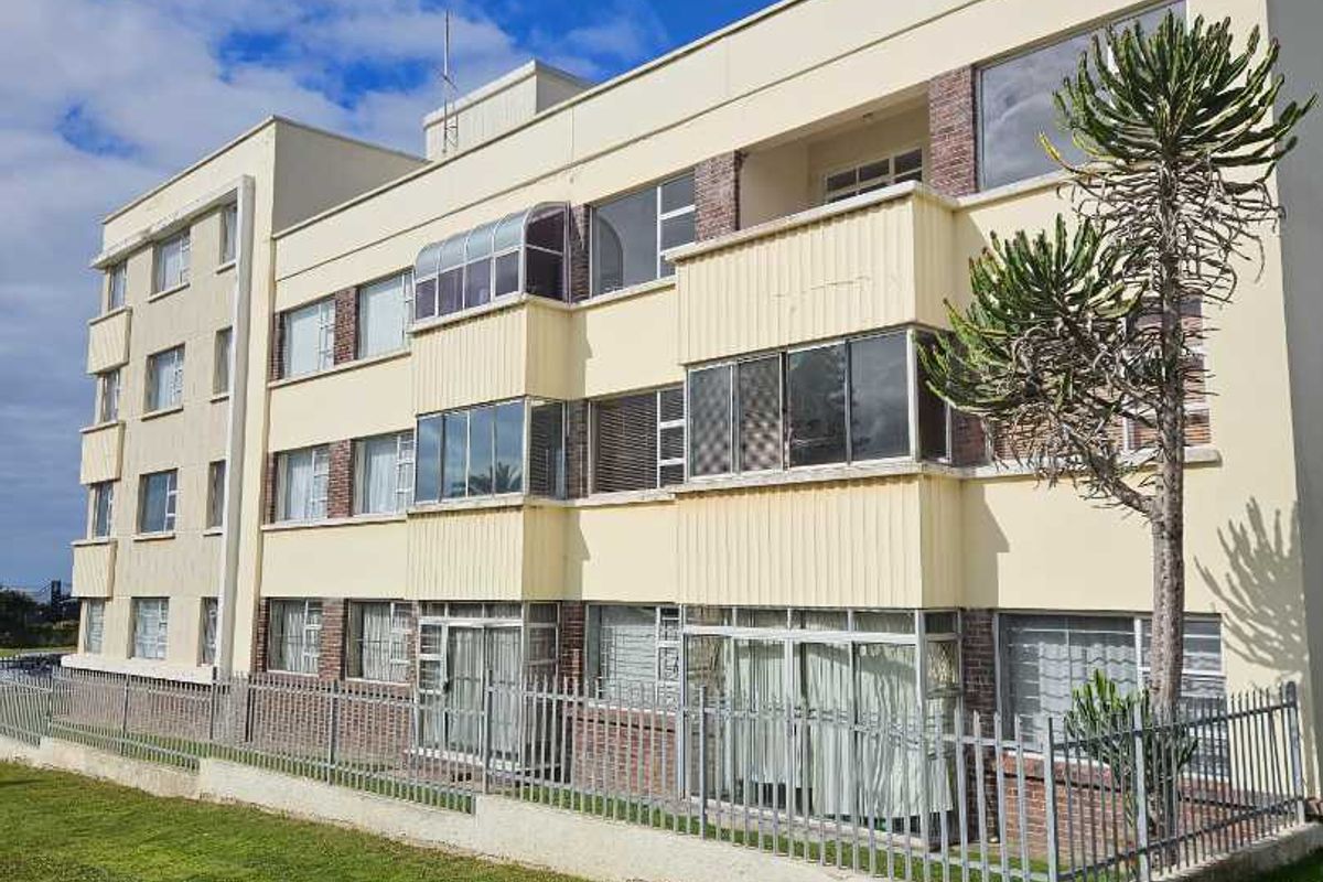 Exterior view of a multi-story apartment building featuring enclosed balconies, brick masonry base, and perimeter metal security fencing set against a lawn.