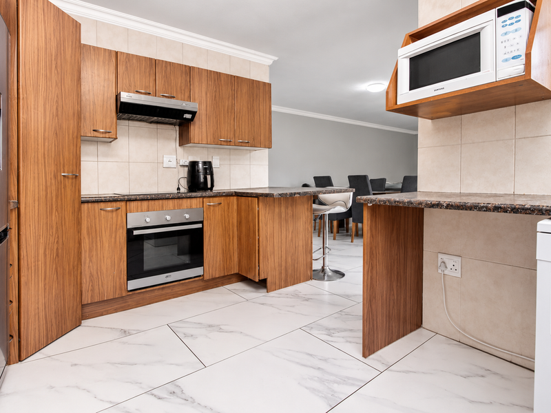 Functional kitchen featuring wood-finish cabinetry, granite countertops, built-in oven, extractor fan, and modern marble-look floor tiles, seamlessly flowing into the dining area.