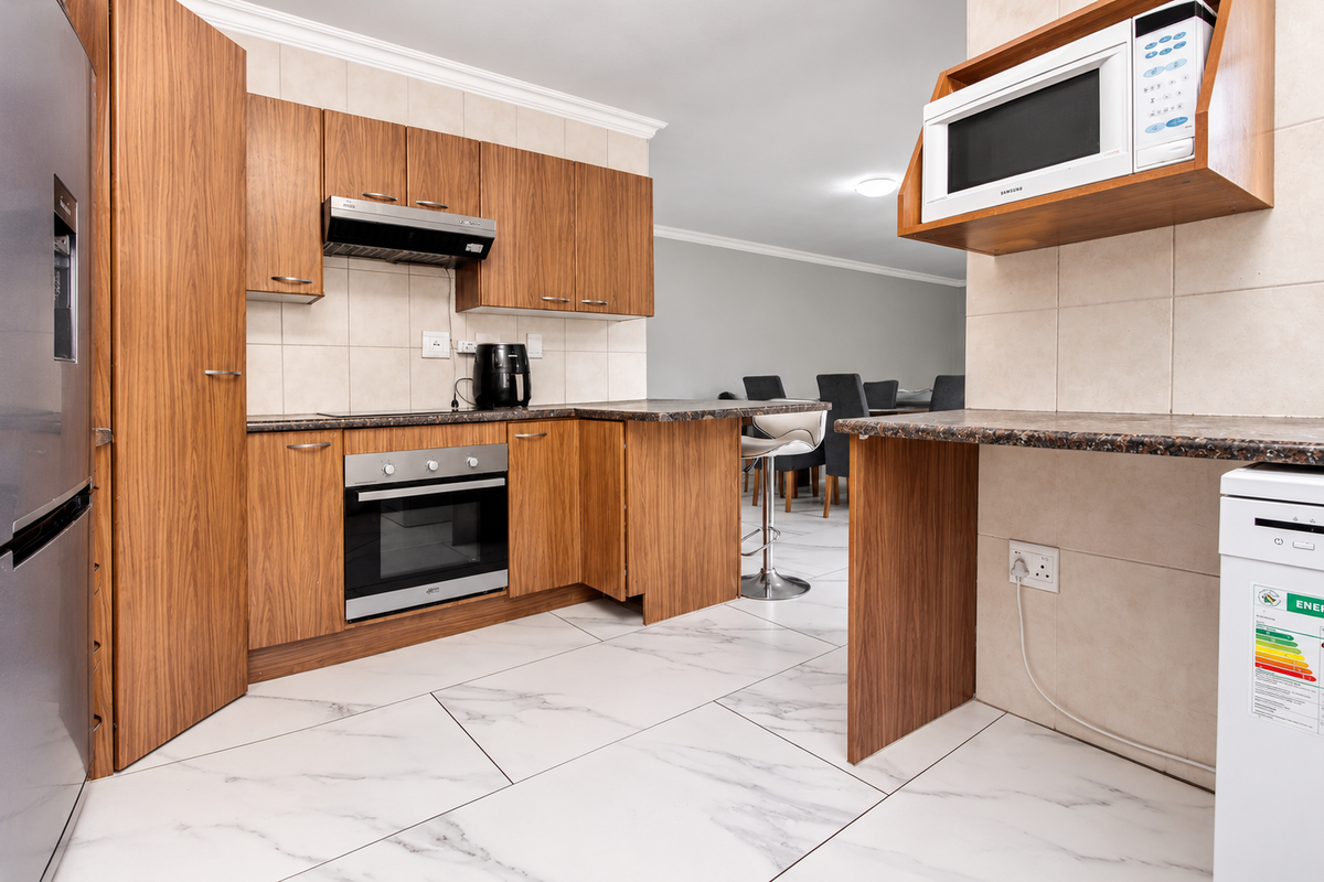 Functional kitchen featuring wood-finish cabinetry, granite countertops, built-in oven, extractor fan, and modern marble-look floor tiles, seamlessly flowing into the dining area.