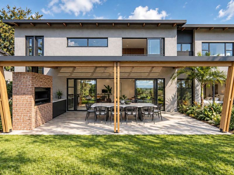 A beautifully designed rear exterior featuring a covered patio with a timber pergola, built-in brick fireplace, outdoor kitchen area, and seamless indoor-outdoor flow.