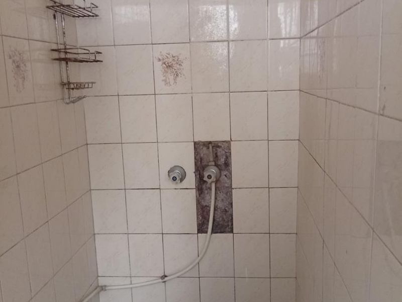 A shower enclosure featuring white wall tiles and dark mosaic floor tiles, currently showing signs of wear and damaged tiling around the shower mixer.