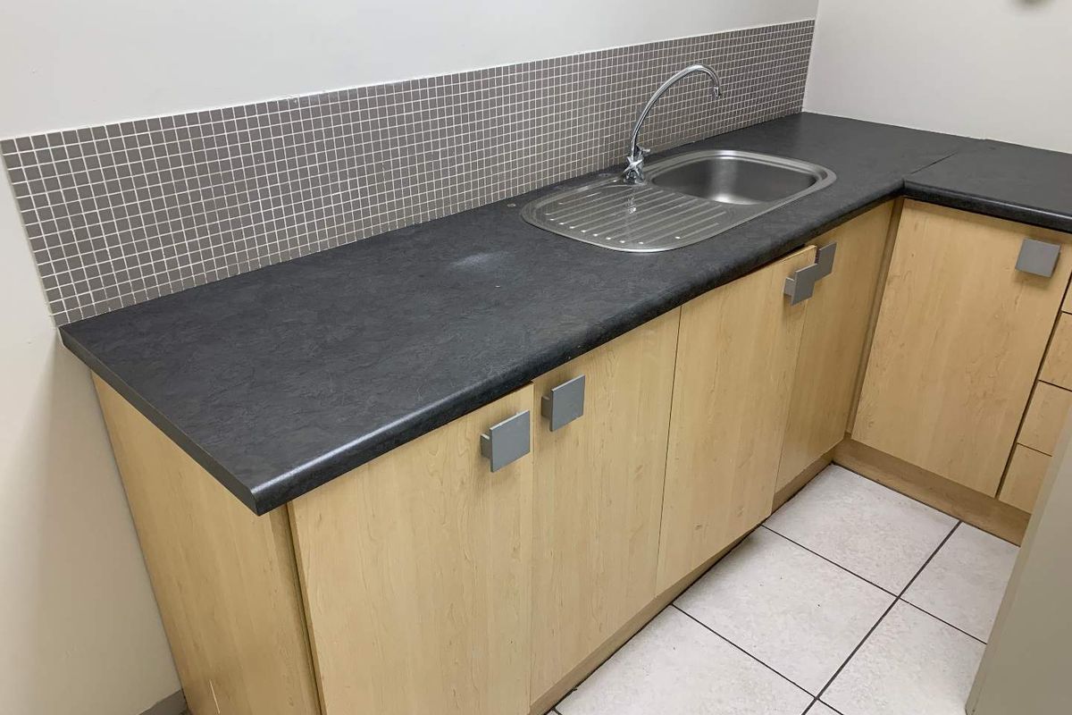 A functional office kitchenette featuring light wood cabinetry, a dark laminate countertop, stainless steel sink, and a mosaic tile backsplash.