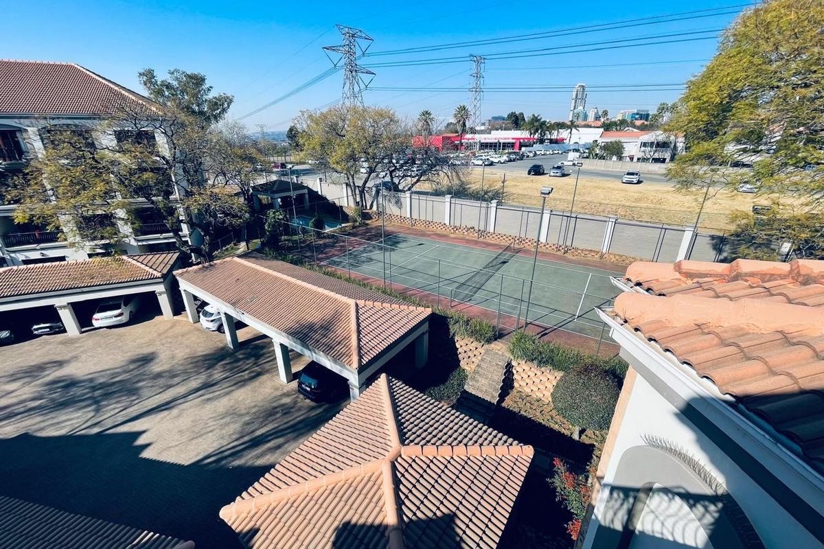 Elevated view of the apartment complex grounds, featuring a tennis court, covered parking bays, and surrounding perimeter fencing.