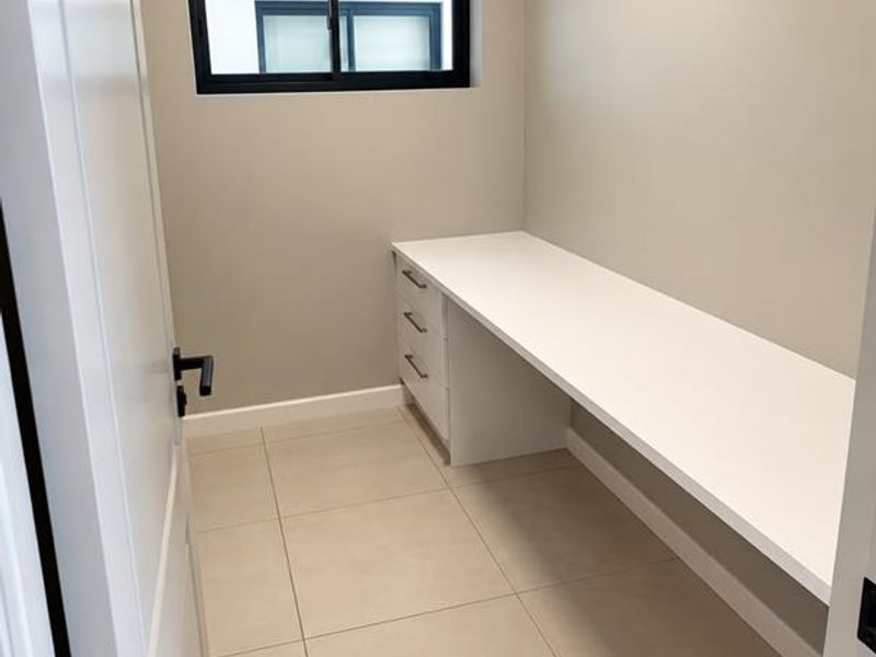 A clean, modern home office featuring neutral wall tones, large format floor tiles, and a functional built-in desk with storage drawers.