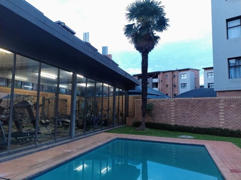 Communal pool area featuring a paved surround, brick boundary wall, and adjacent glass-fronted gym facility within the apartment complex.
