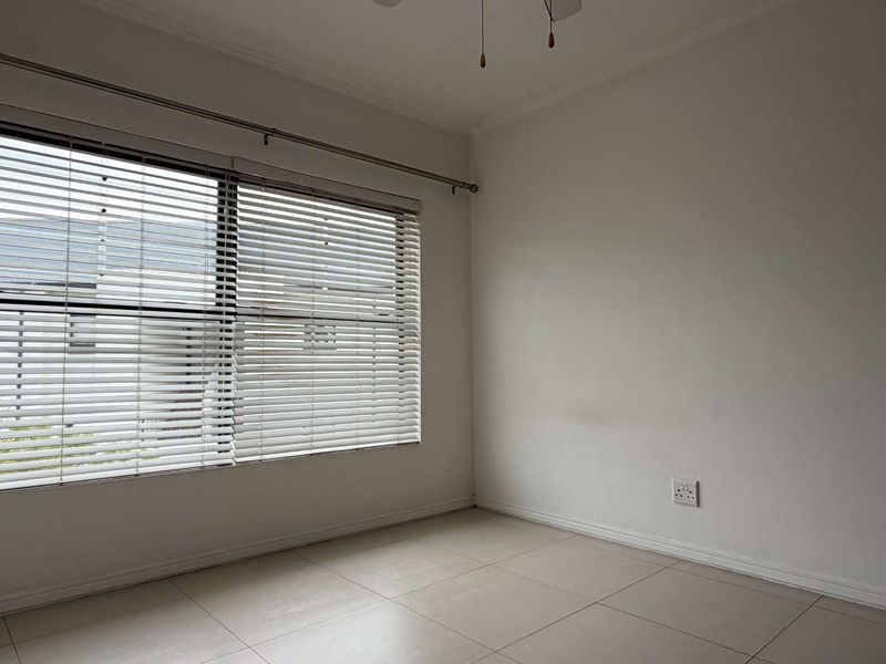 A bright, empty bedroom featuring neutral tiled flooring, a ceiling fan, and venetian blinds on the window.