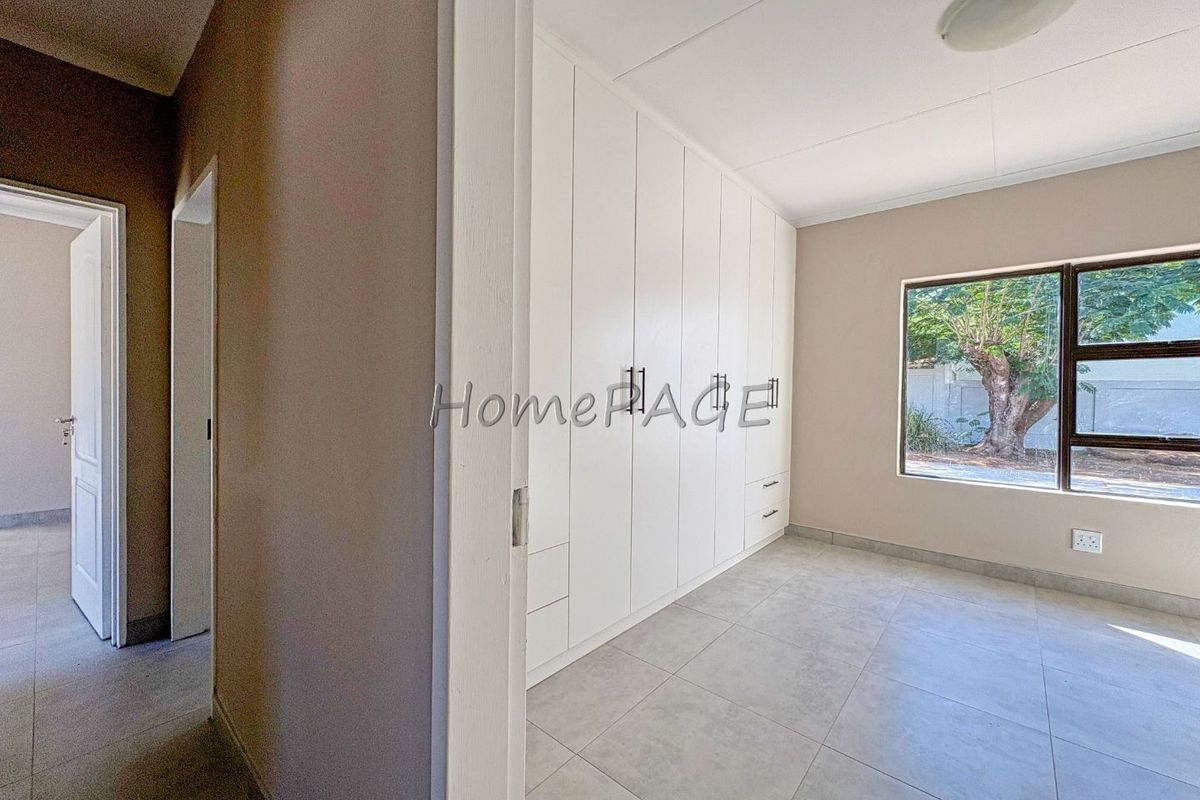 A bright, empty bedroom featuring modern tiled flooring, neutral walls, and extensive white built-in cupboards with sleek hardware.