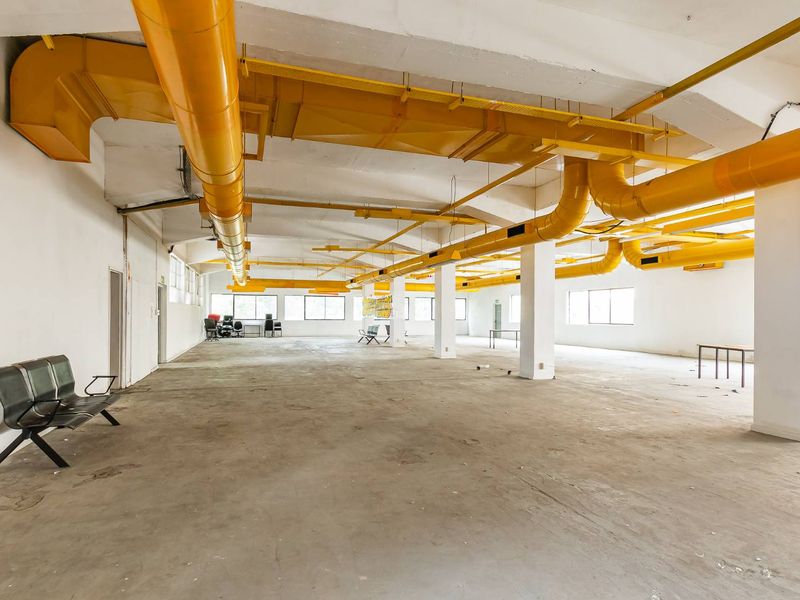 Expansive open-plan industrial space featuring exposed yellow HVAC ducting, concrete flooring, and large windows, offering a versatile layout for various commercial uses.