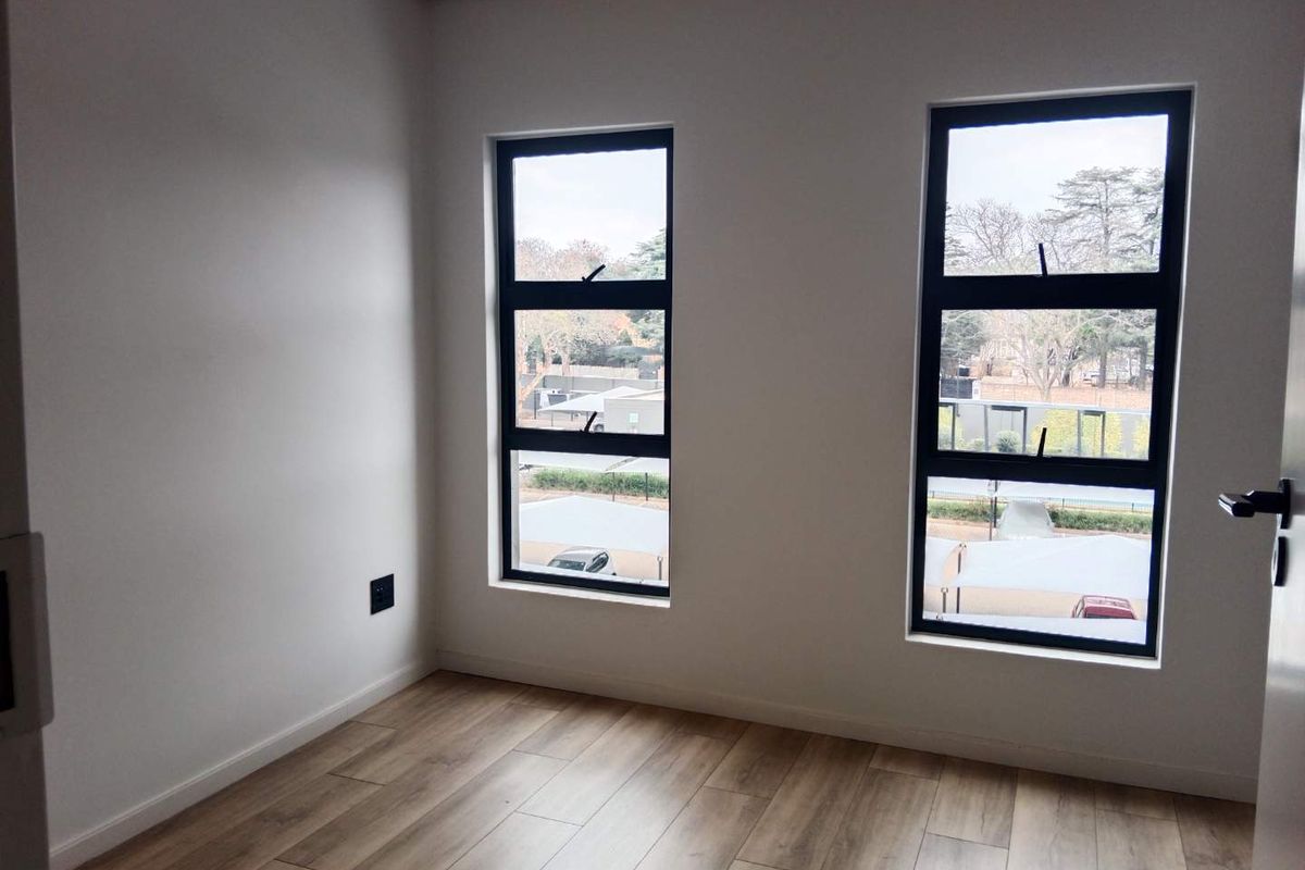 A bright, empty bedroom featuring modern laminate flooring, neutral walls, and large aluminium-framed windows providing natural light.