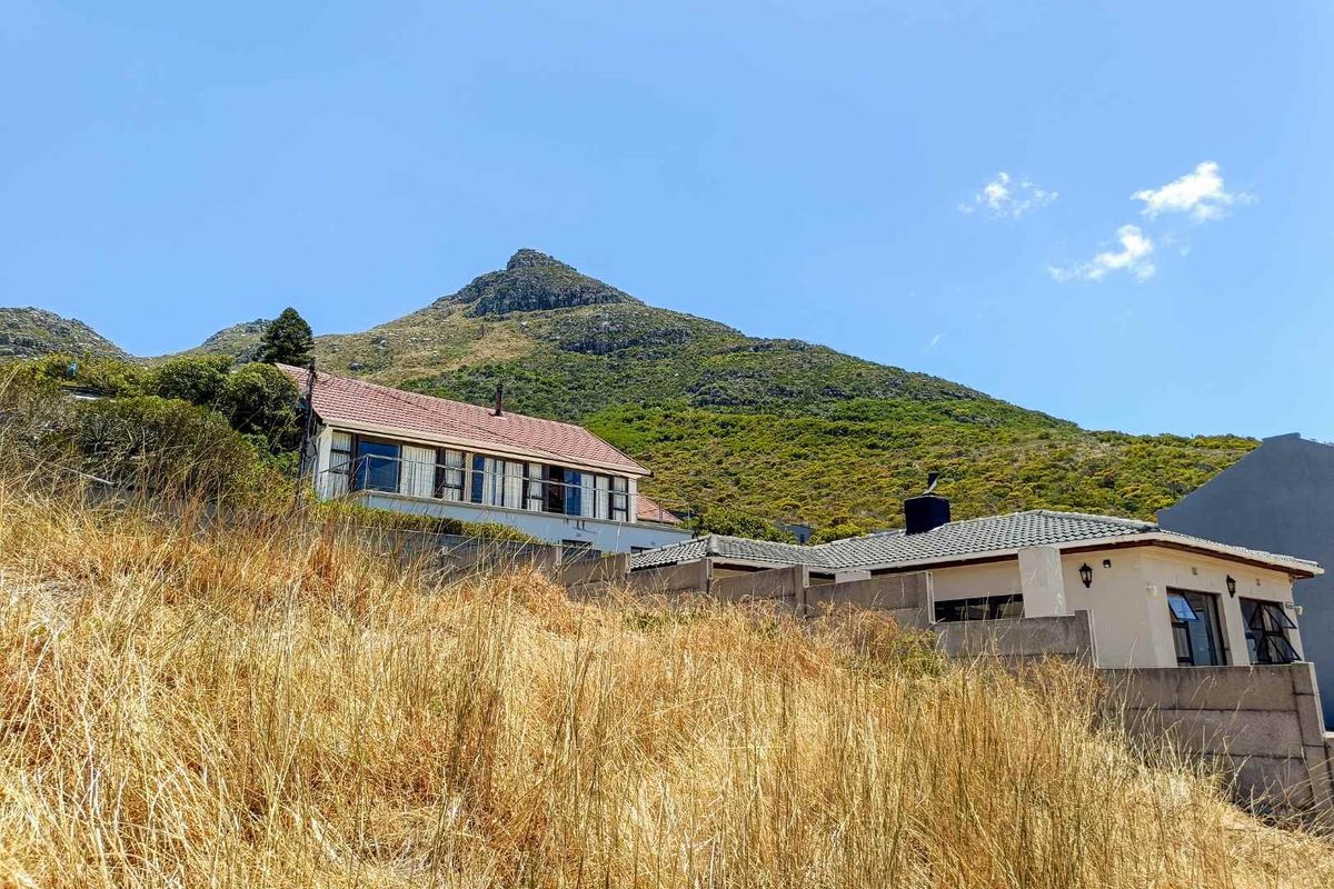 A view of the residential property set against a mountain backdrop, featuring a sloped plot with dry grass and established building structures.