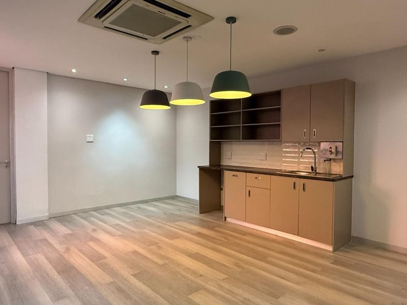 A clean, modern office kitchenette featuring built-in cabinetry, a sink, tiled backsplash, and contemporary pendant lighting, set within a bright, open-plan workspace.