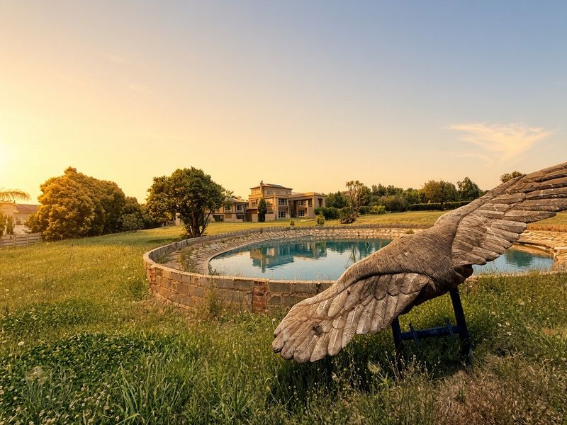 An exterior view of a large residential property featuring a stone-walled swimming pool set within expansive, overgrown grounds at sunset.
