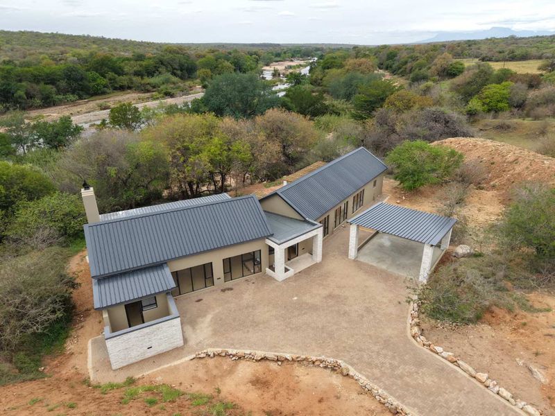 Aerial view of a modern house with a metal roof, stone accents, and a carport, set in a scenic natural landscape.