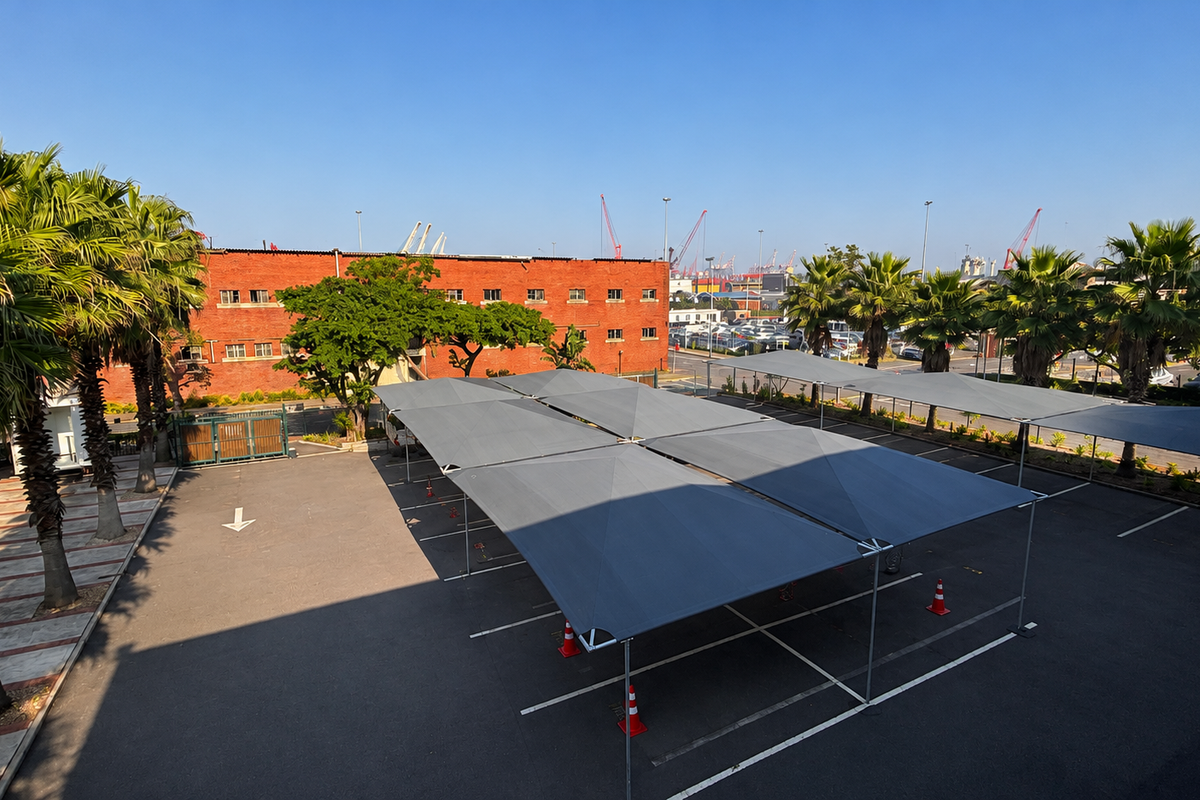 An elevated view of a paved office parking area featuring multiple shade cloth carports, mature palm trees, and a red brick building in the background.