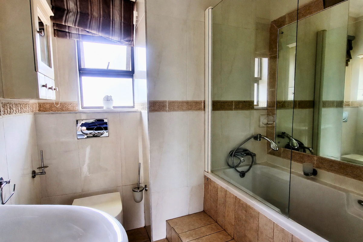 A well-maintained bathroom featuring a built-in vanity, wall-mounted toilet, and a bathtub with a glass shower screen and tiled finishes.