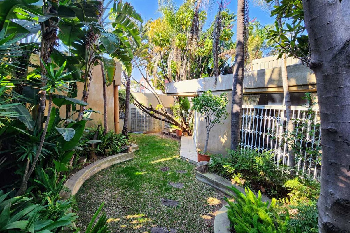 A private rear garden featuring curved concrete edging, established tropical landscaping, a small tiled patio area, and secure access to the home.
