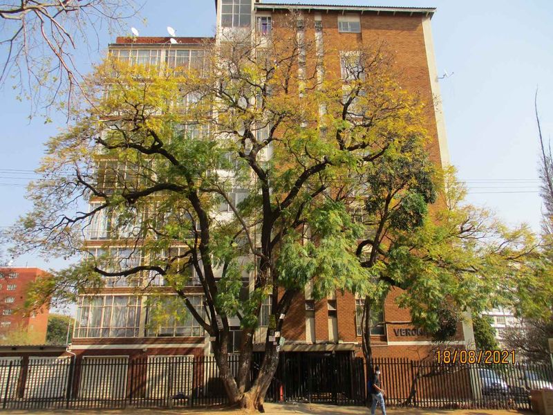 Front elevation of a multi-story apartment building featuring a brick facade, enclosed balconies, and secure perimeter fencing.