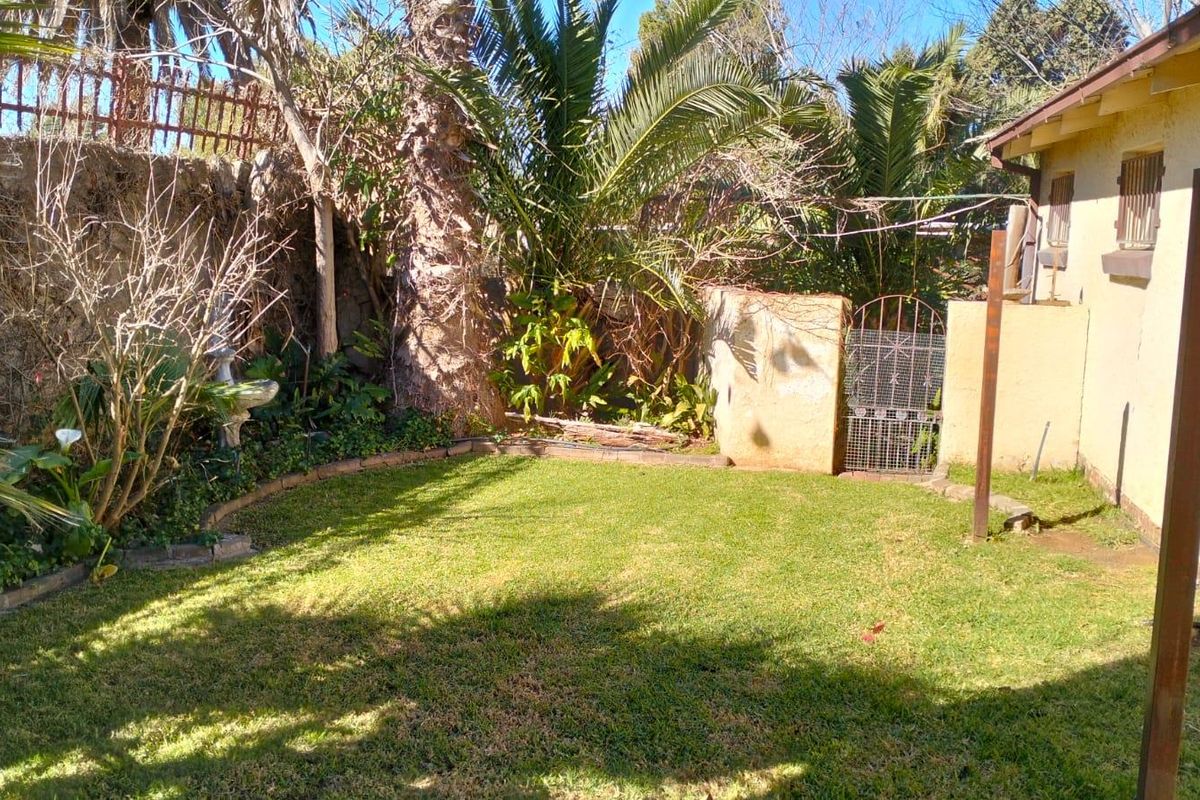 A private rear garden area featuring a lawn, established palm trees, and a metal security gate, adjacent to the side of the house.