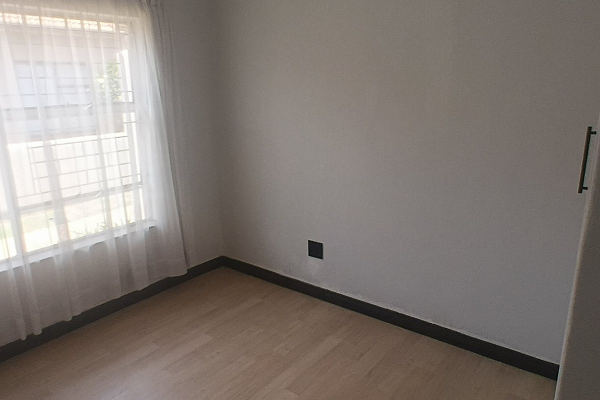 A bright, empty bedroom featuring light laminate flooring, a built-in cupboard, and a window fitted with security bars.