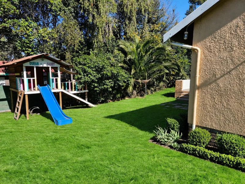 A well-maintained residential garden featuring a lush green lawn, mature trees, and established shrubbery alongside the side elevation of the house.