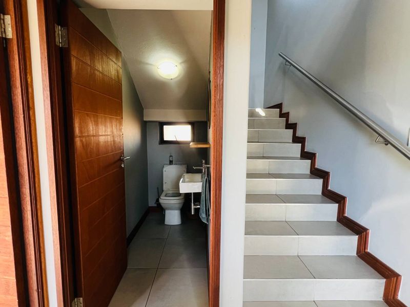 A compact guest bathroom located under the staircase, featuring a wall-mounted basin and tiled flooring, adjacent to a tiled staircase with a metal handrail.