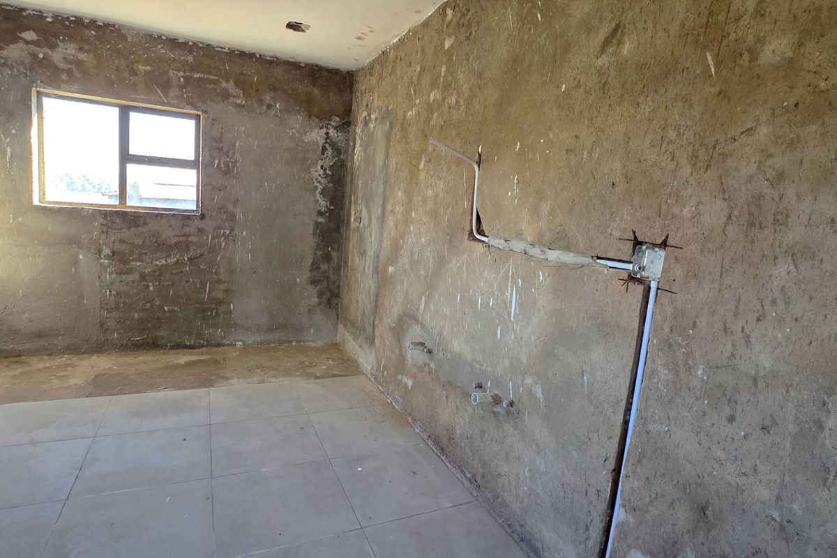 An unfinished kitchen space featuring tiled flooring, exposed plumbing, and electrical conduits, requiring significant renovation and wall finishing to become functional.