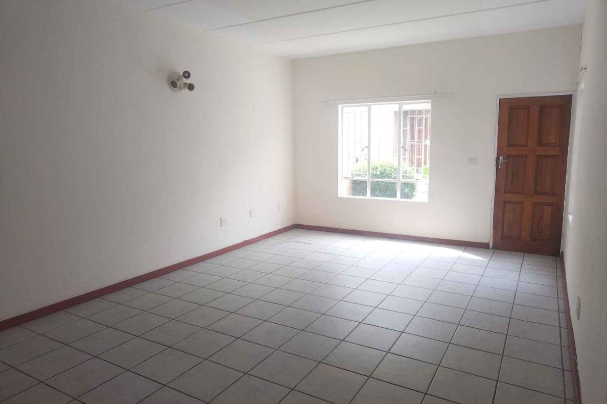 A spacious, empty living area featuring neutral tiled flooring, a large window with security bars, and a solid wood entrance door.