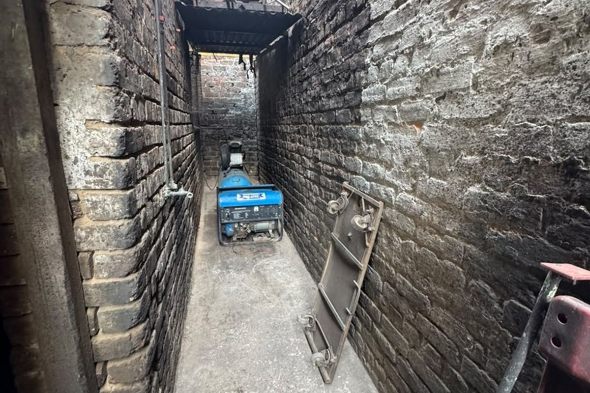 A narrow exterior alleyway featuring exposed brick walls and concrete flooring, showing significant signs of wear and weathering.