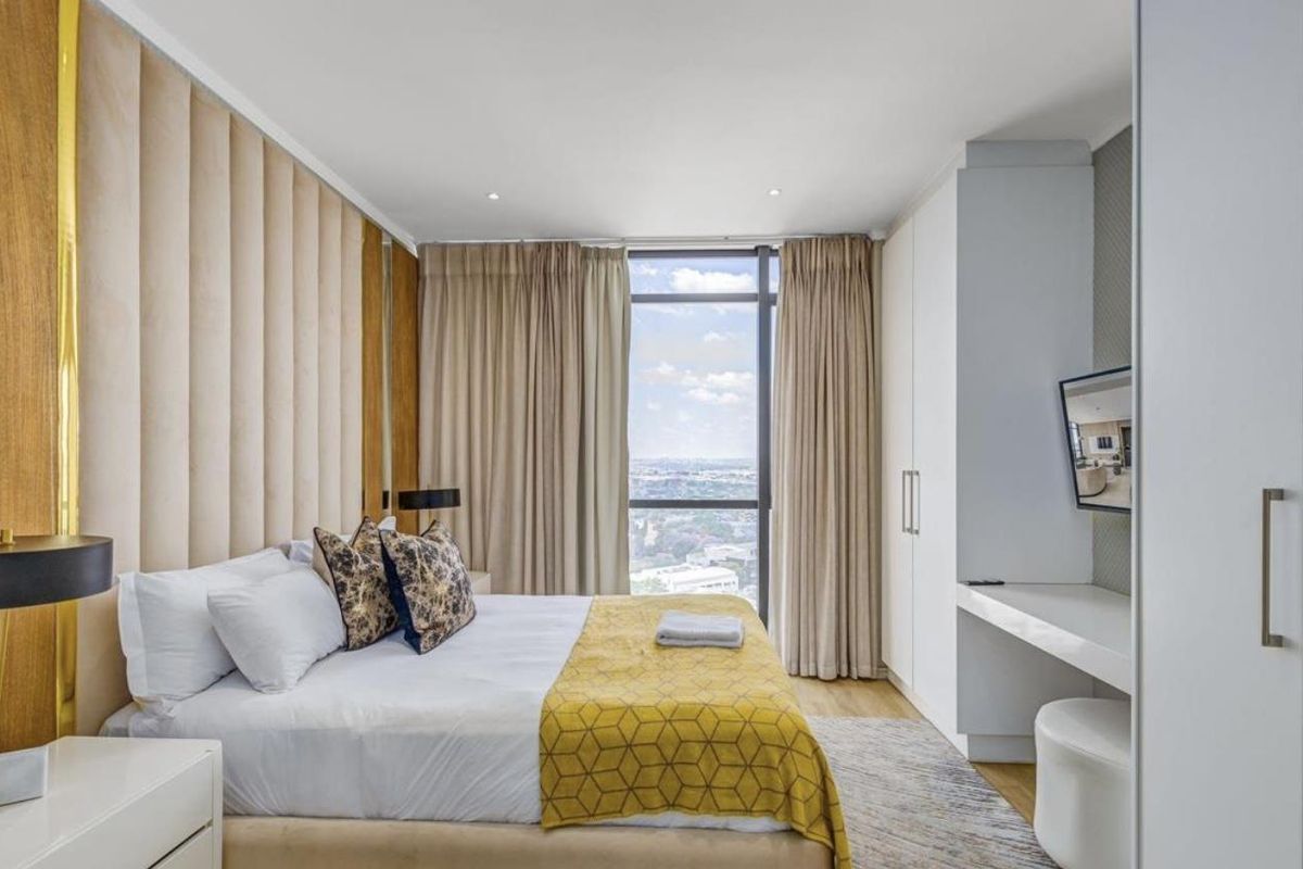 Modern bedroom featuring a floor-to-ceiling window with city views, custom upholstered headboard wall, and built-in cabinetry with integrated desk space.