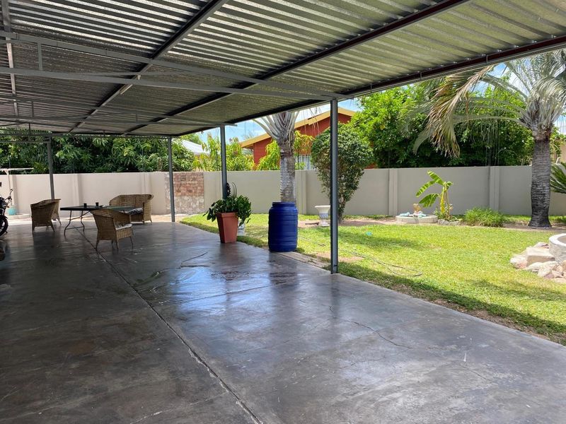 A spacious covered patio area featuring a corrugated metal roof, steel support structure, and concrete flooring, overlooking a private garden with boundary walls.
