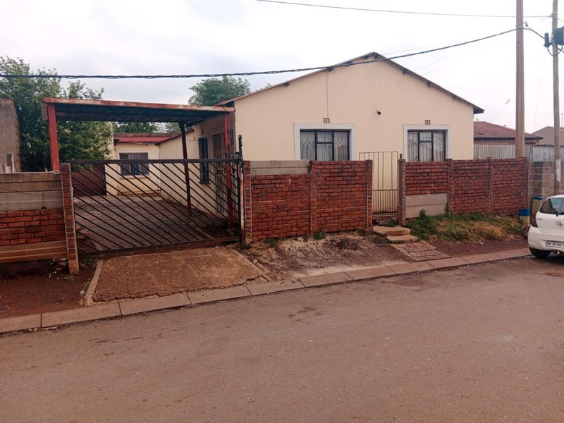 Front exterior view of a single-story house featuring a brick boundary wall, metal driveway gate, and a covered carport area.