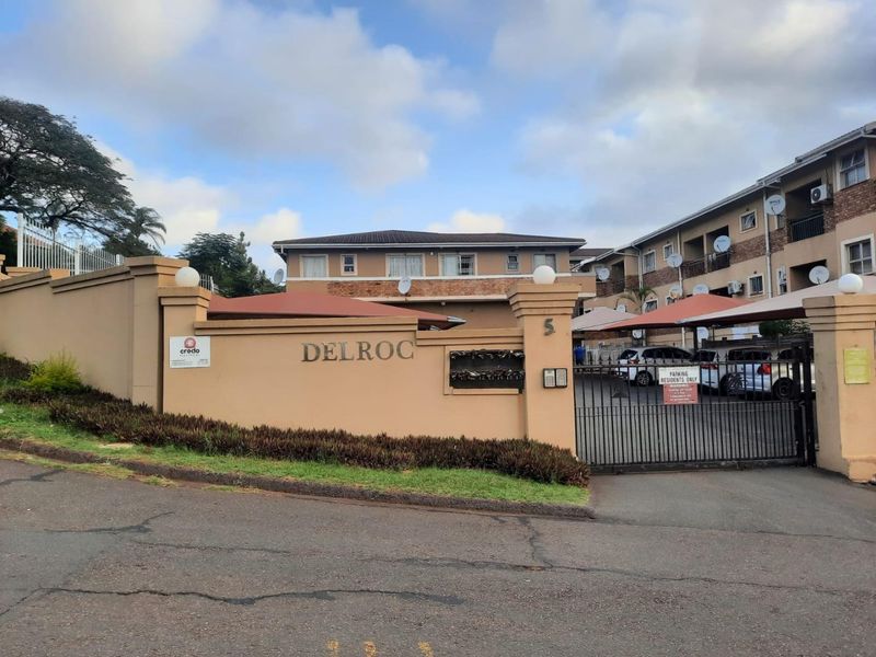 Front entrance view of Delroc apartment complex featuring a secure gated access point, perimeter wall, and covered parking bays for residents.