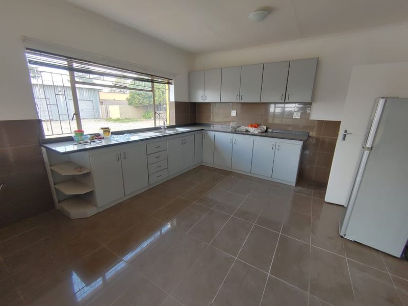 A functional, empty kitchen featuring ample built-in cabinetry, tiled flooring, a stainless steel sink, and a large window with security bars.