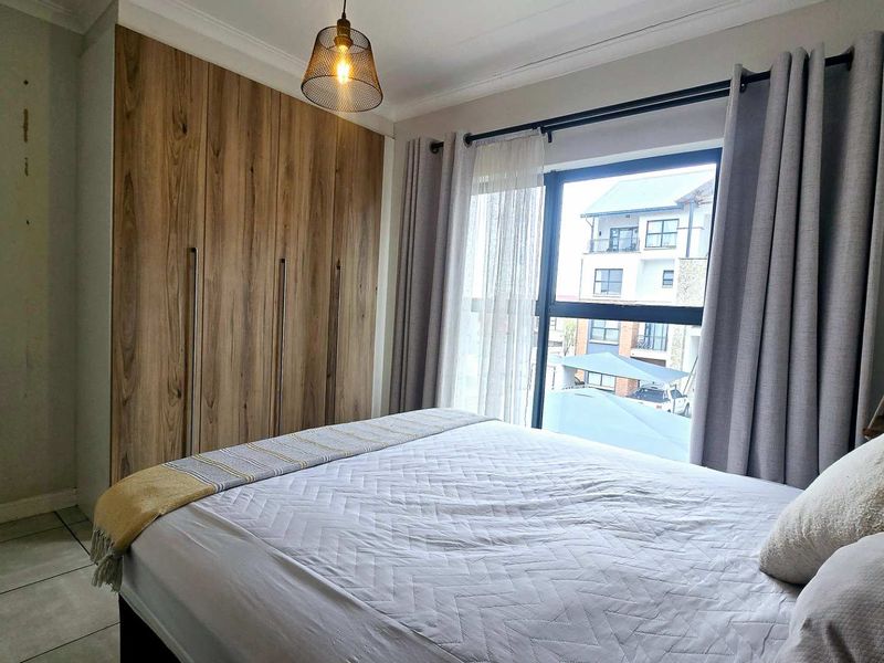 A well-lit bedroom featuring modern wood-finish built-in cupboards, tiled flooring, and a large window providing natural light and exterior views.