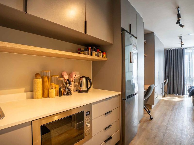 Modern galley-style kitchen featuring sleek grey cabinetry, stone countertops, integrated appliances, and efficient track lighting, seamlessly flowing into the adjacent living area.