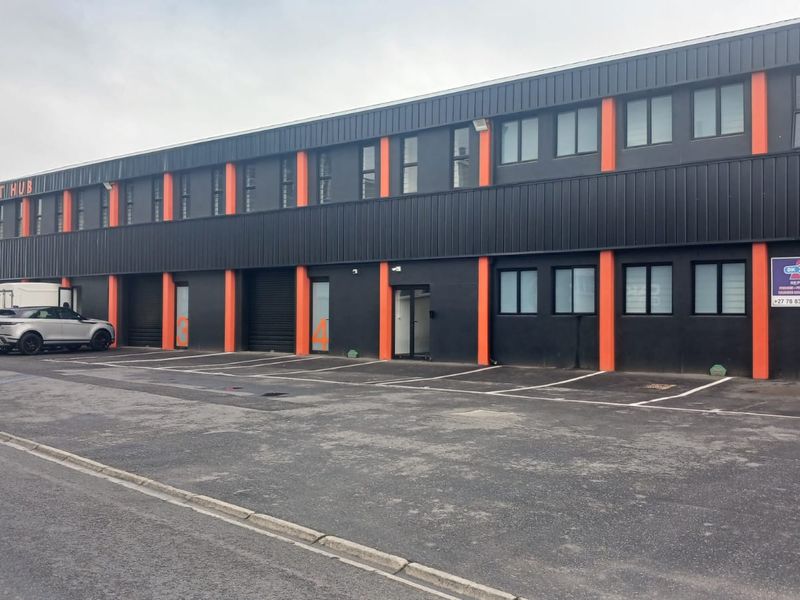Modern industrial commercial building featuring a dark metal facade with orange accents, multiple roller shutter doors, and dedicated asphalt parking bays.