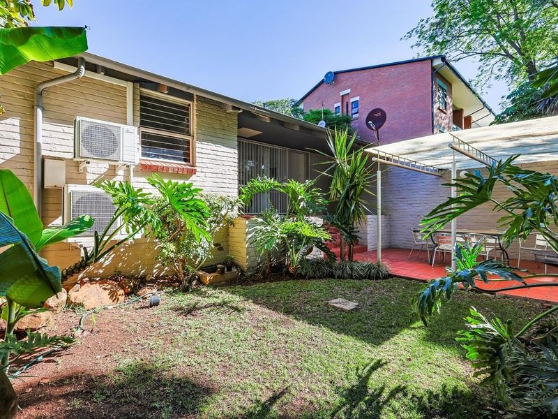 A private rear courtyard featuring a paved patio area, established garden, and exterior brick facade with mounted air conditioning units.