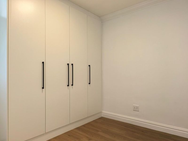 A clean, modern bedroom featuring white built-in cupboards, light wood-look laminate flooring, and classic coving detail.