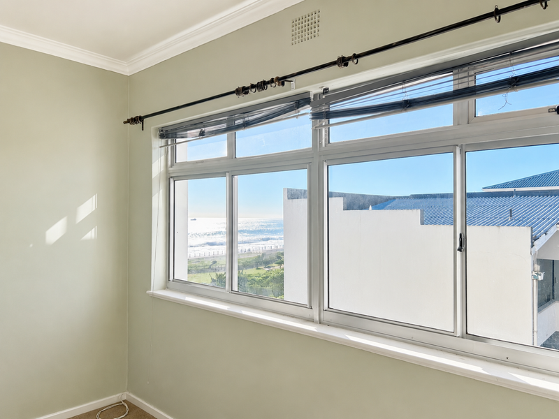 A bright, empty bedroom featuring large aluminium-framed windows with ocean views, neutral wall finishes, and classic cornice detailing.