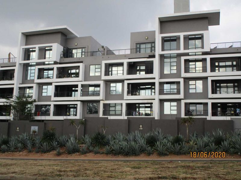 A modern multi-story apartment complex featuring a contemporary grey and white facade with private balconies and secure perimeter fencing.