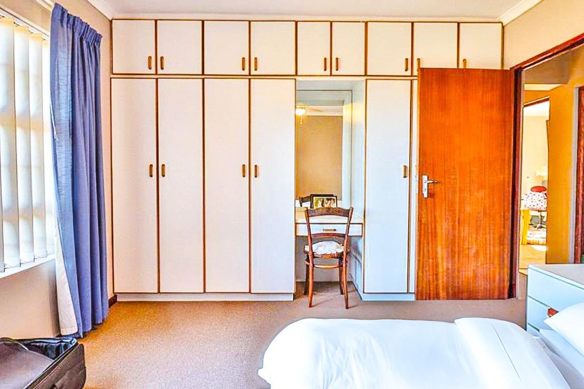 A bedroom featuring extensive built-in cupboards with an integrated vanity station, carpeted flooring, and natural light through vertical blinds.