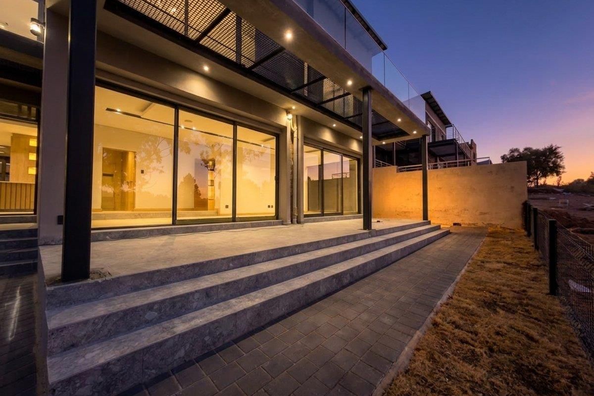 A modern rear exterior view featuring a spacious covered patio with stone steps, glass sliding doors, and a sleek glass balustrade at dusk.
