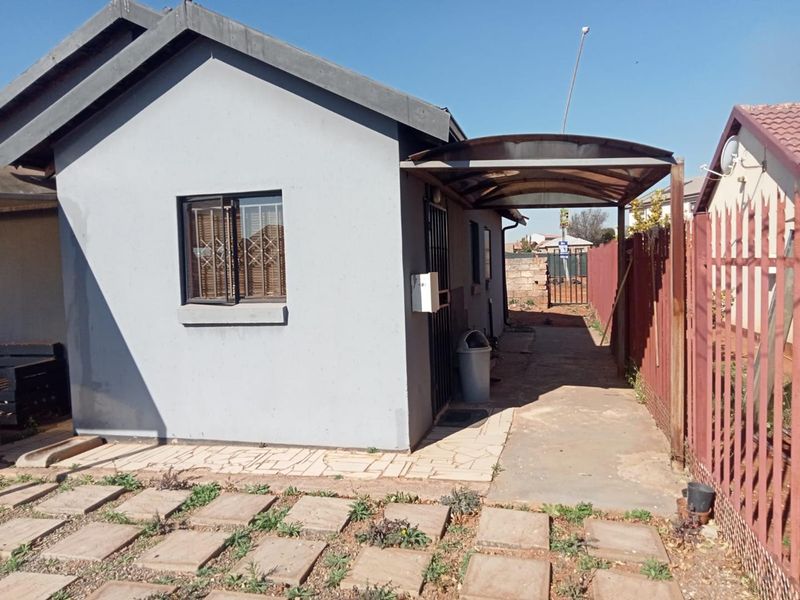 Front exterior view of the house featuring a paved driveway, a covered carport, and metal perimeter security fencing.