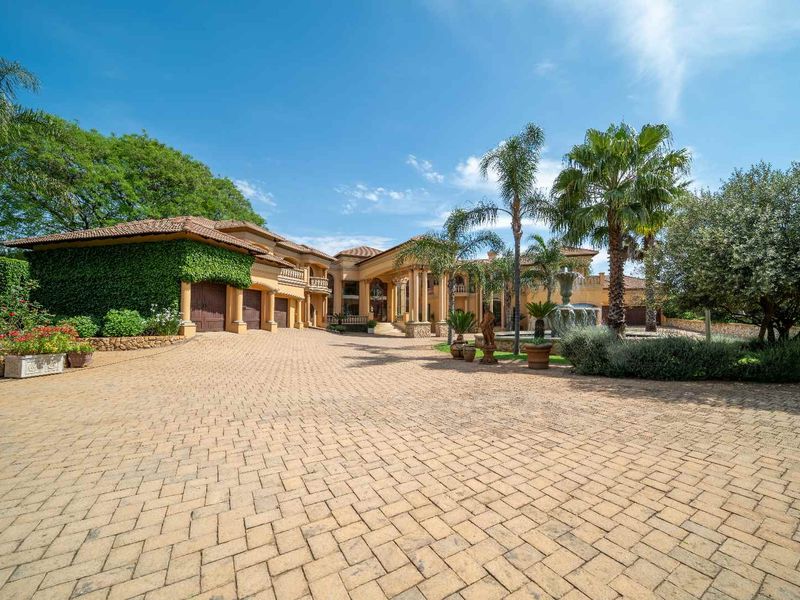 Grand front elevation of a luxury small holding featuring a spacious paved driveway, stone fountain, and impressive architectural columns at the main entrance.