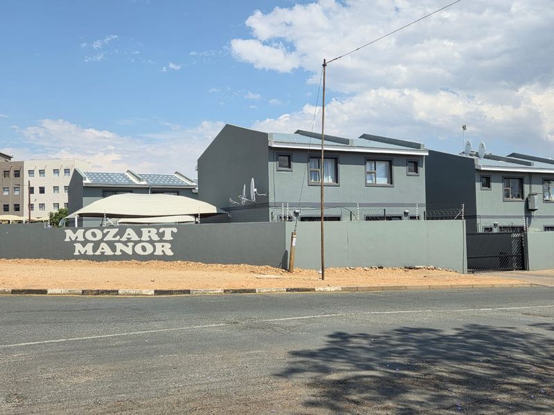 Street view of Mozart Manor townhouse complex featuring grey painted facades, perimeter wall with electric fencing, and covered parking structures.