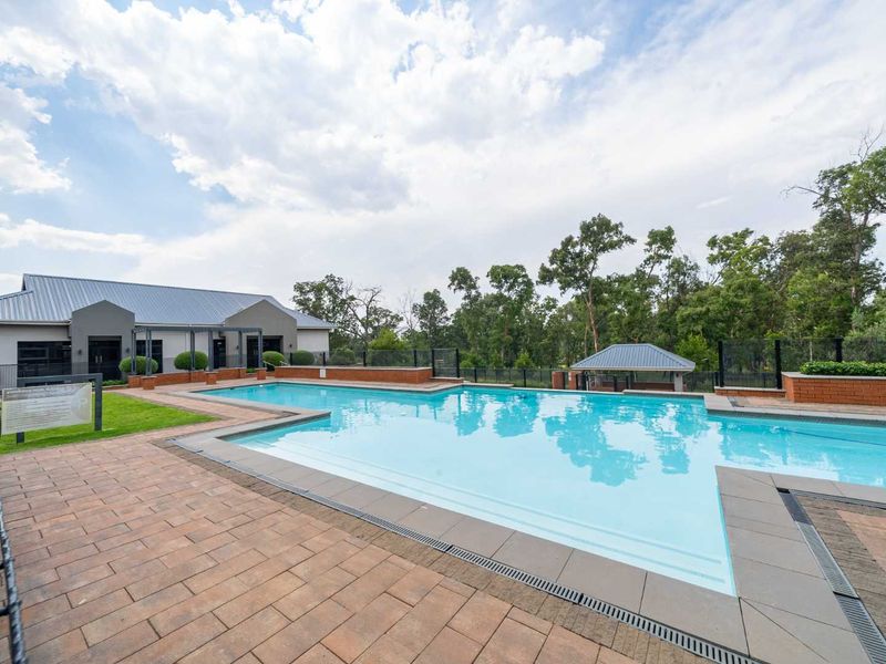 A well-maintained communal swimming pool area featuring brick paving, glass fencing, and a surrounding landscaped garden with a covered structure.