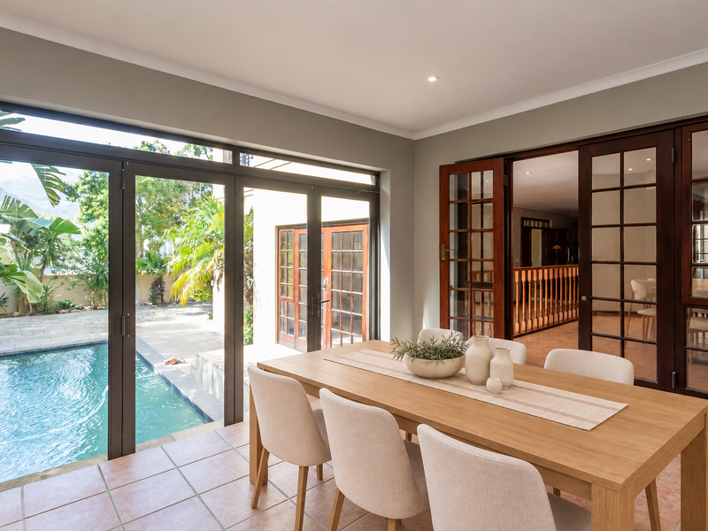 A bright, covered patio area featuring tiled flooring, large aluminium sliding doors opening to a swimming pool, and wooden french doors leading indoors.