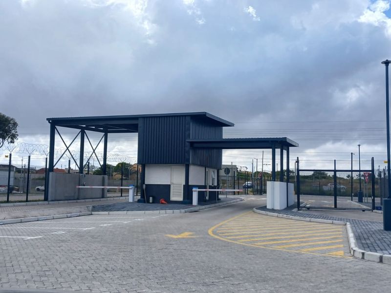 A modern office park entrance featuring a security guard house, boom gates, and paved access lanes with perimeter security fencing.
