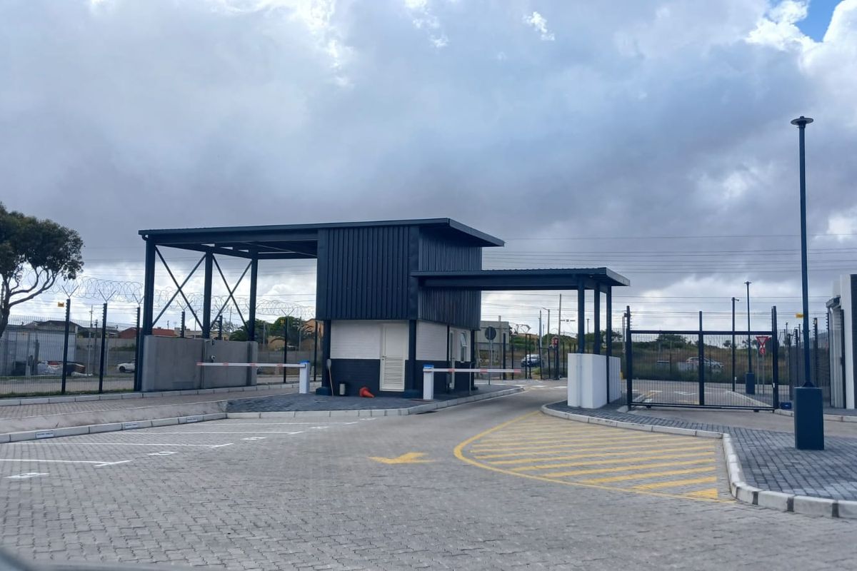 A modern office park entrance featuring a security guard house, boom gates, and paved access lanes with perimeter security fencing.