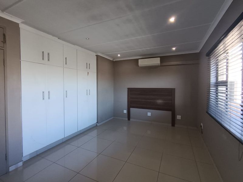 A spacious bedroom featuring neutral tiled flooring, extensive built-in cupboards, a wall-mounted air conditioning unit, and venetian blinds.